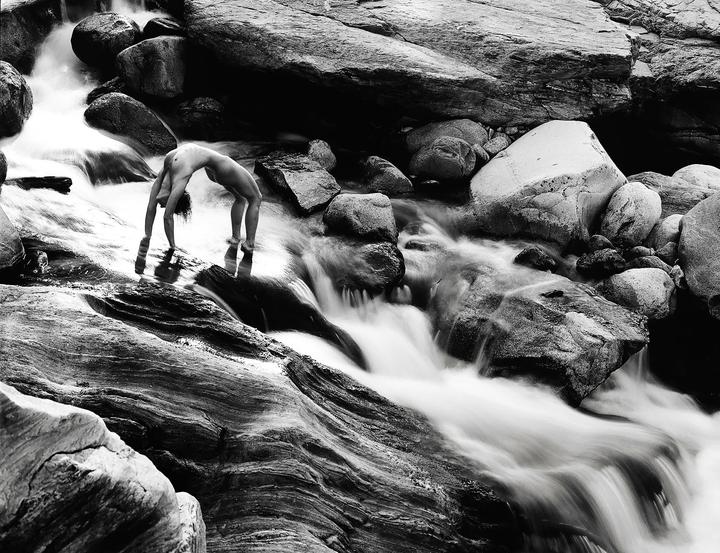 A nude woman performs a backbend in a rocky river with flowing water.