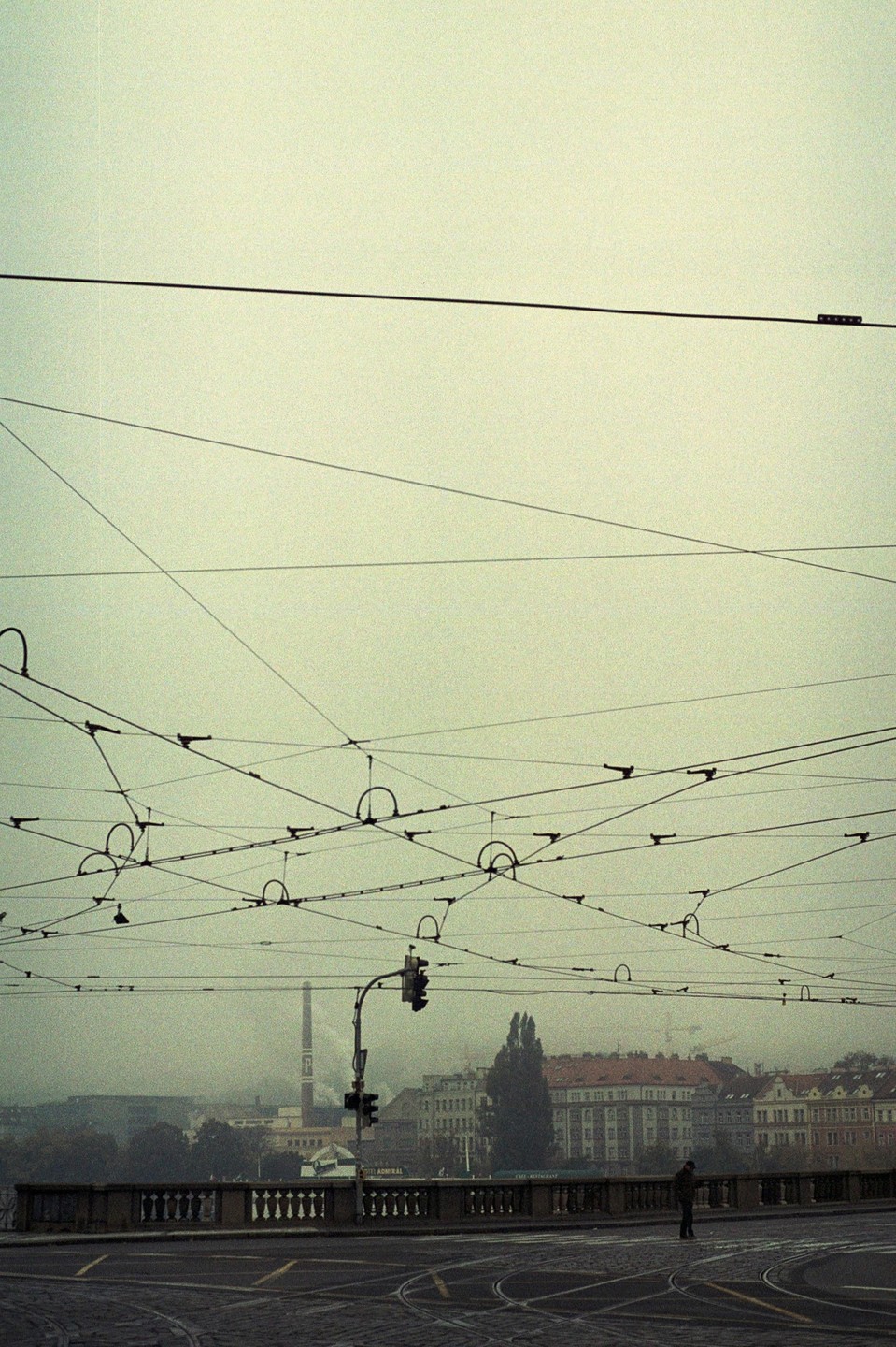 A man stands on a bridge with many overhead wires and buildings in the background on a hazy day.