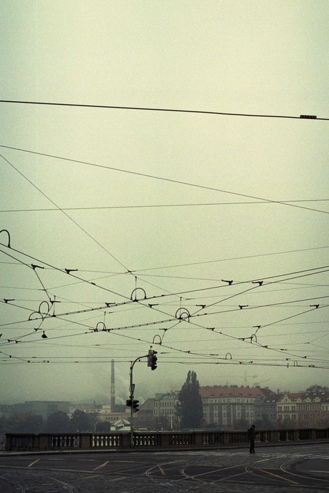 A man stands on a bridge with many overhead wires and buildings in the background on a hazy day.