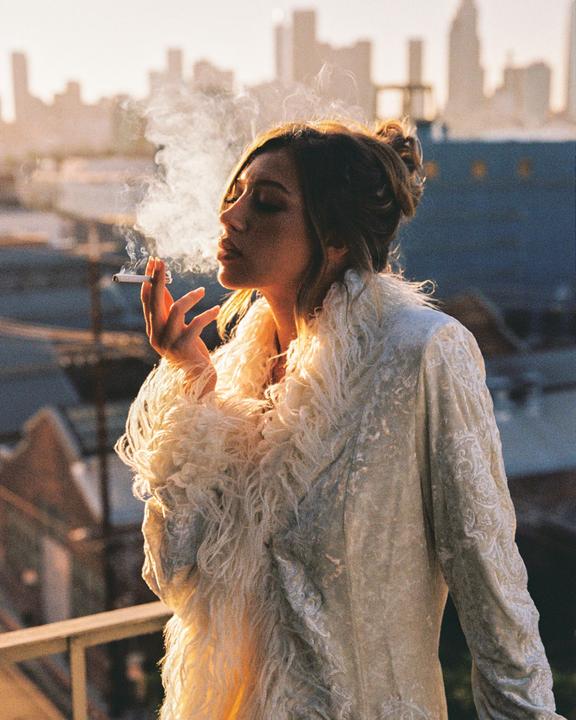 A woman in a fluffy coat smokes a cigarette with a city skyline in the background.