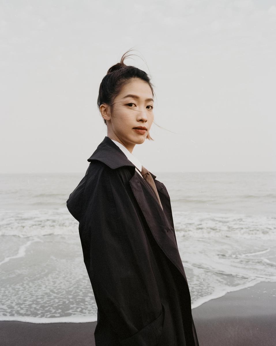 A woman in a dark coat stands on a beach with waves crashing behind her.