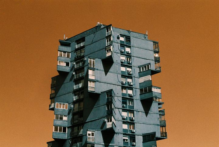 A tall, angular building with many windows and balconies stands against an orange sky.