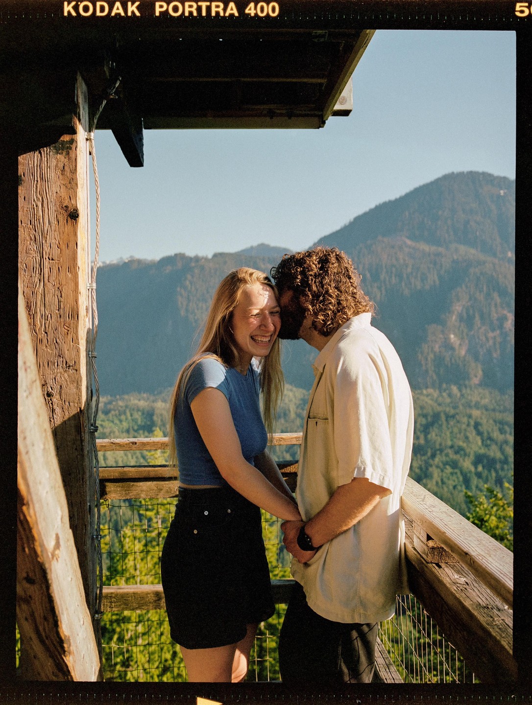 A couple stands on a wooden balcony overlooking a mountain landscape, with the man kissing the woman's cheek as she laughs.