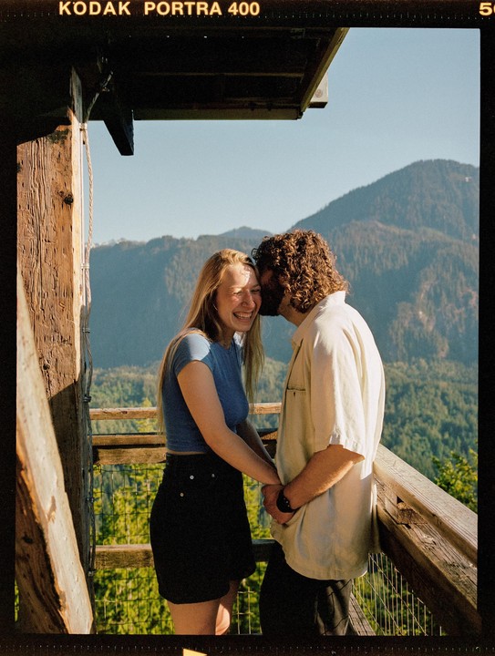 A couple stands on a wooden balcony overlooking a mountain landscape, with the man kissing the woman's cheek as she laughs.