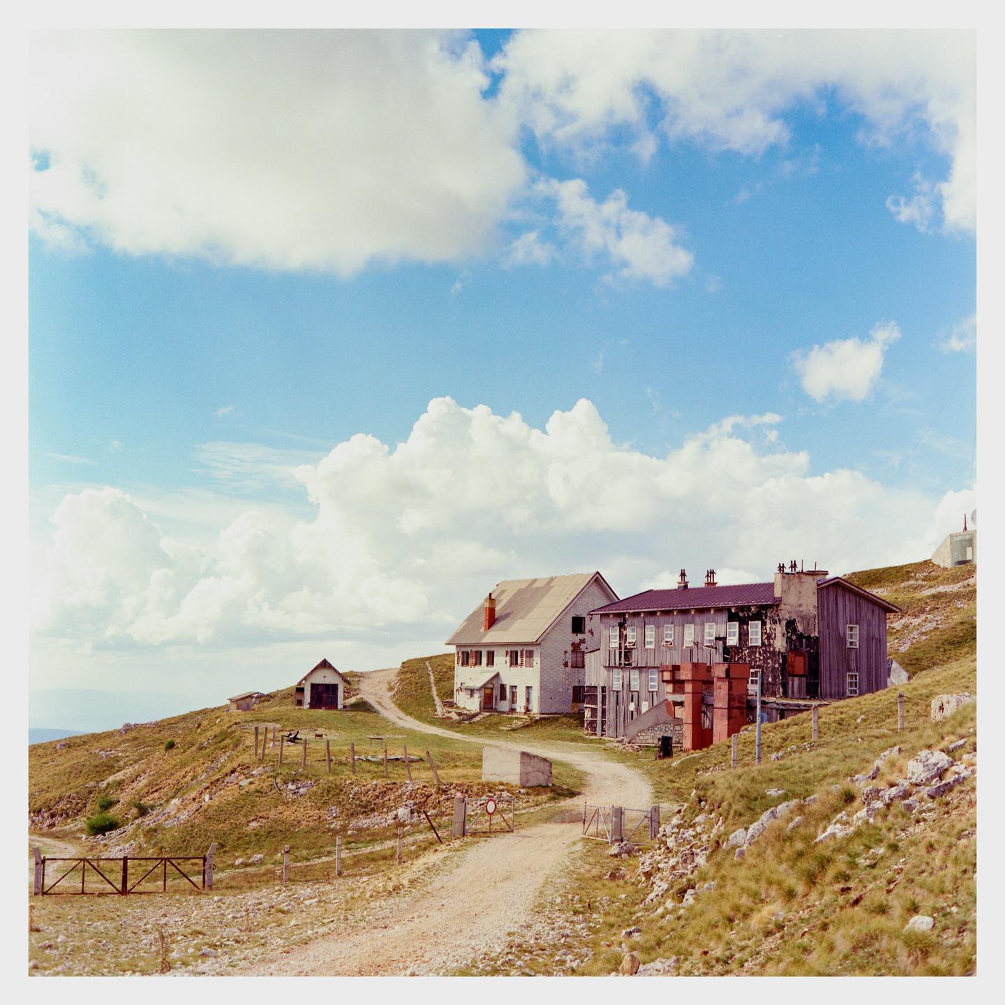 A dirt road winds through a grassy hillside past several buildings under a cloudy sky.