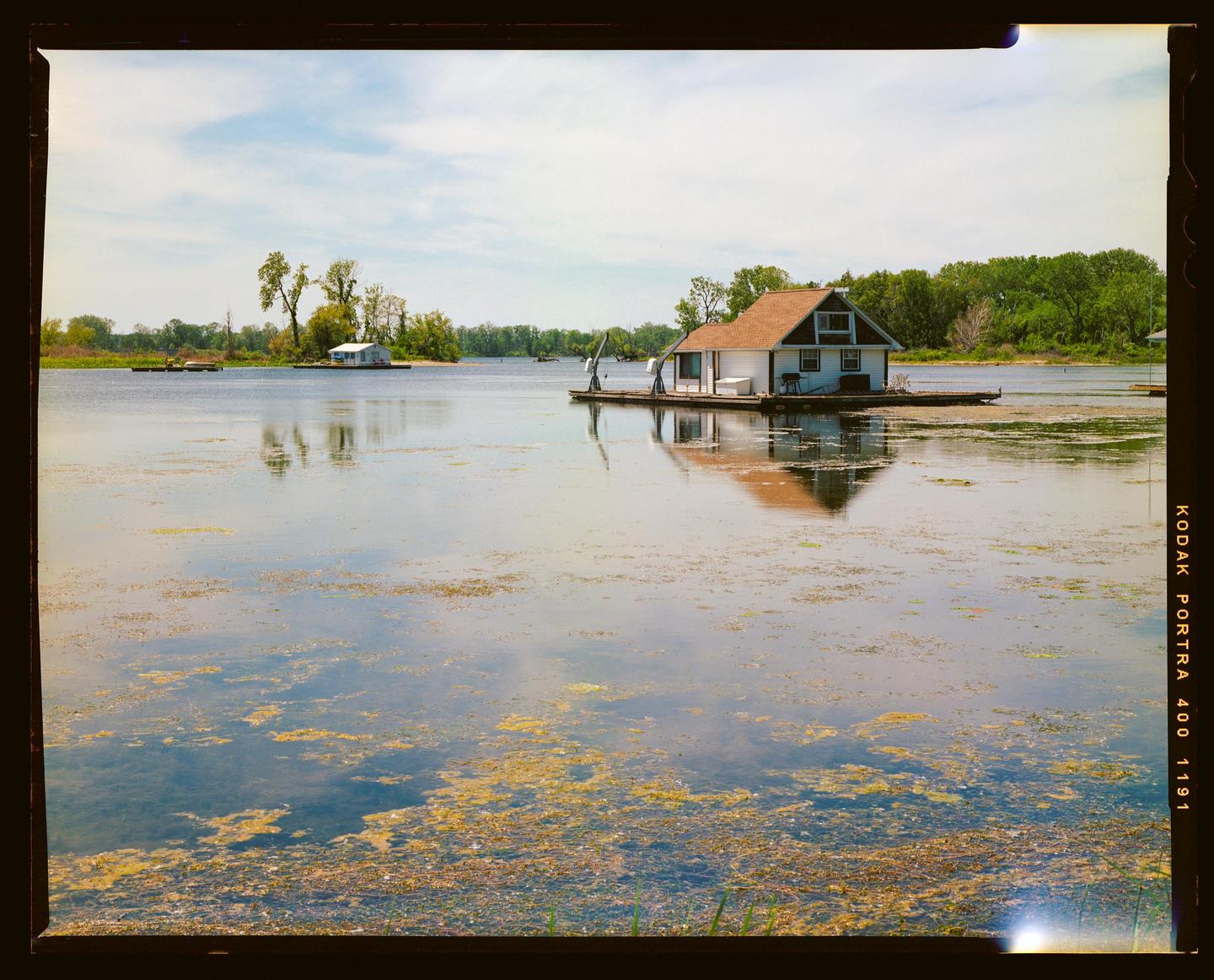 A small houseboat with a brown roof is floating on a lake with green algae and is reflected in the water.
