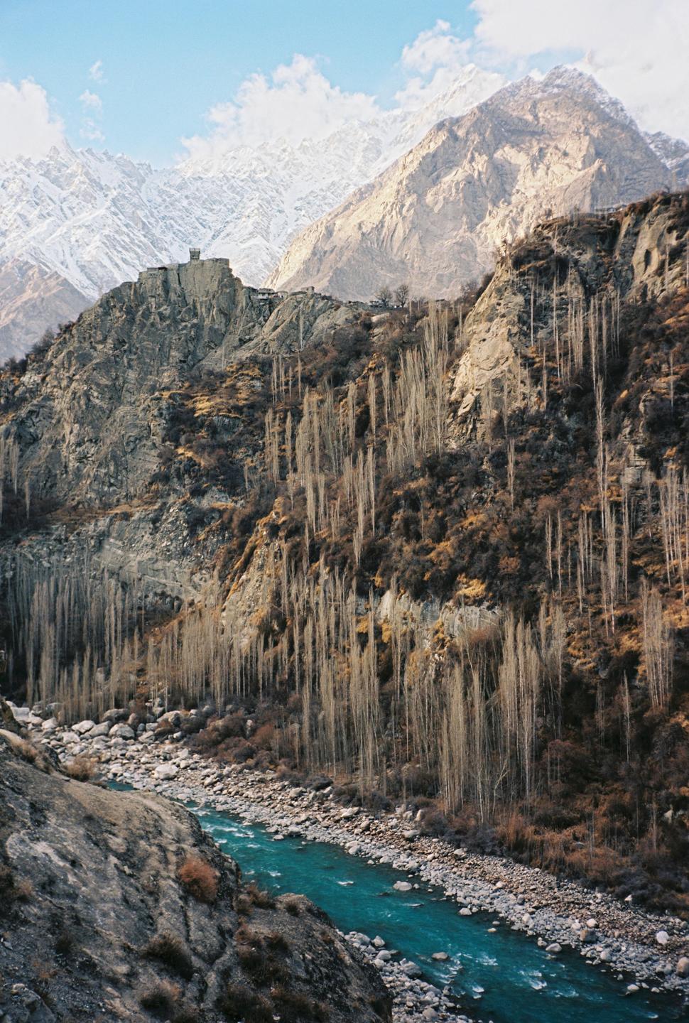 A turquoise river flows through a valley with steep, rocky mountains and a snow-capped peak in the background.