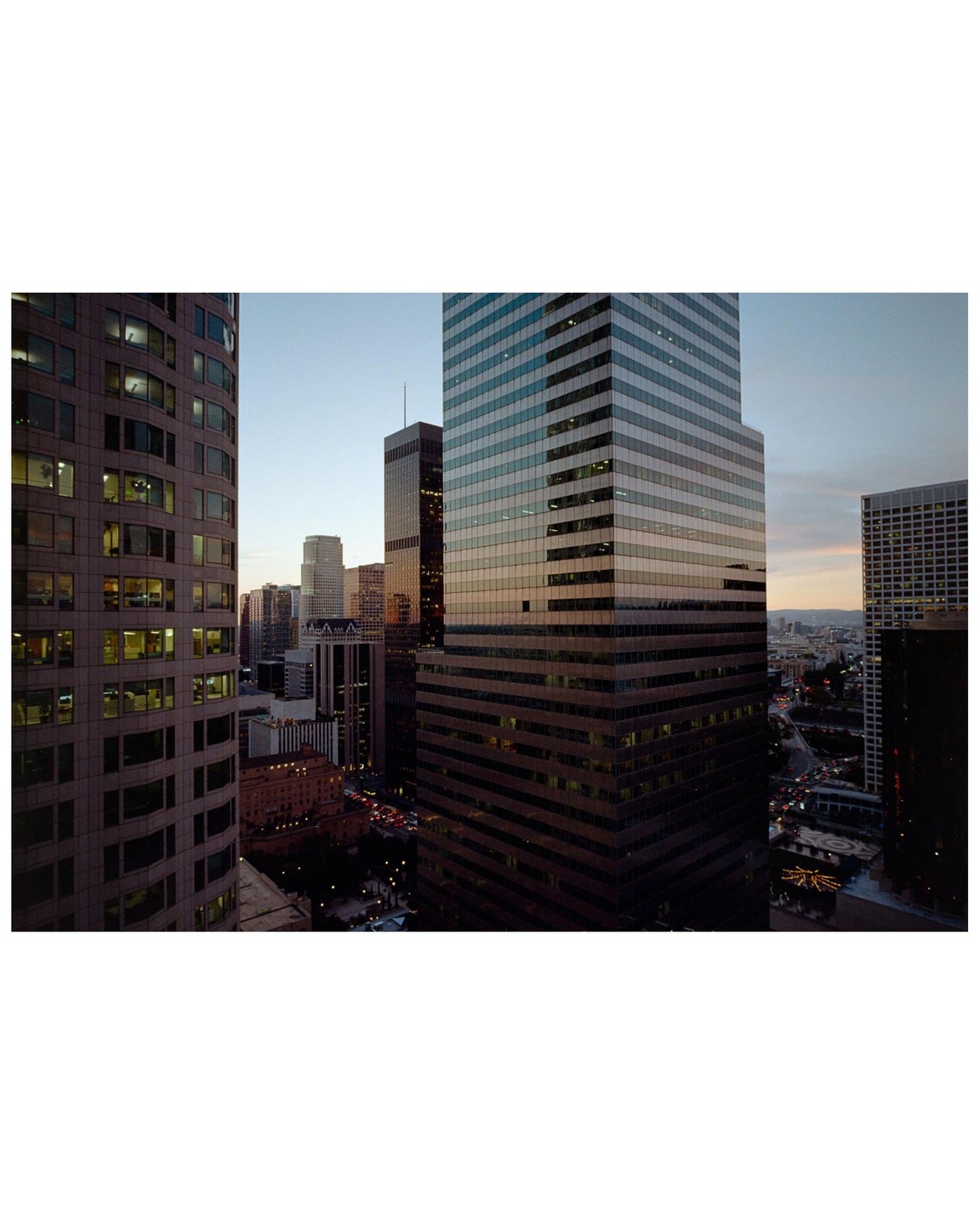 A wide view of a city at dusk shows several tall buildings and a street with car lights.