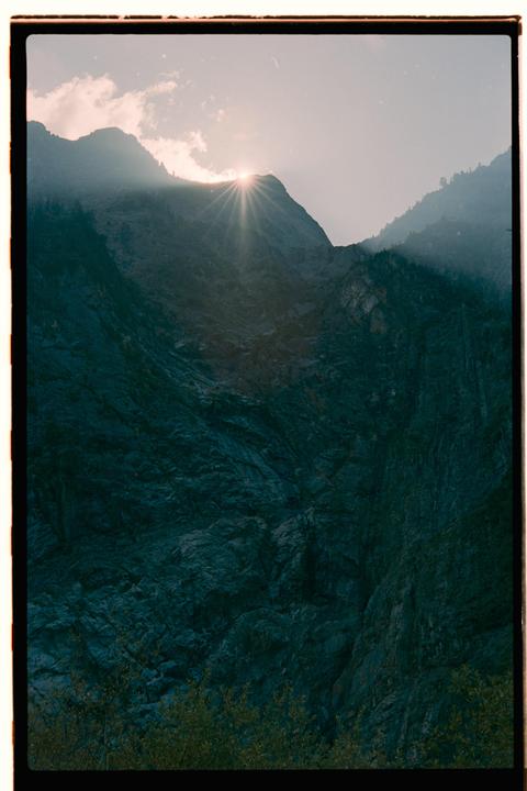 Sunlight bursts over a mountain peak, illuminating the rugged terrain and a hazy sky.