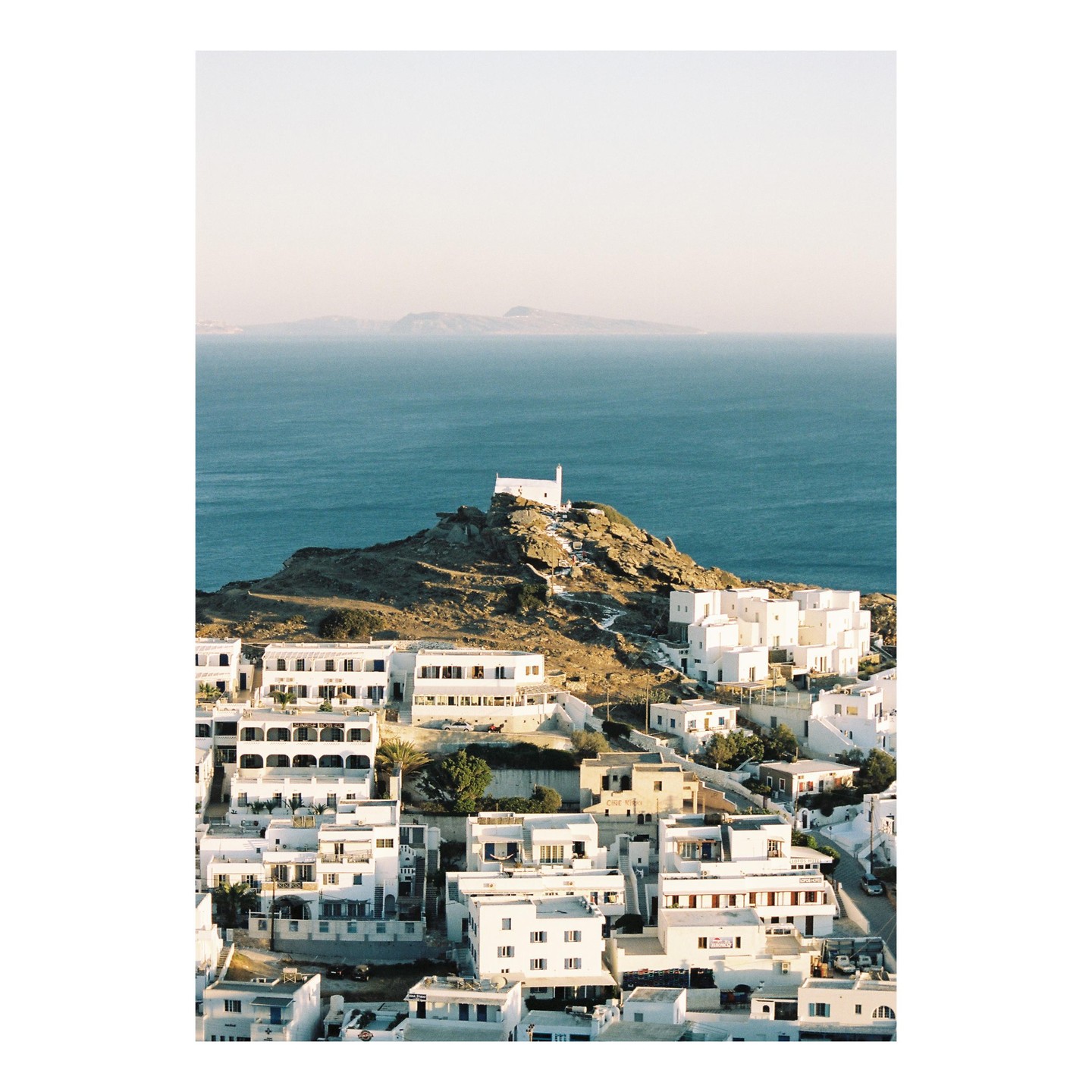 A white church sits atop a rocky hill overlooking a town of white buildings and the sea.