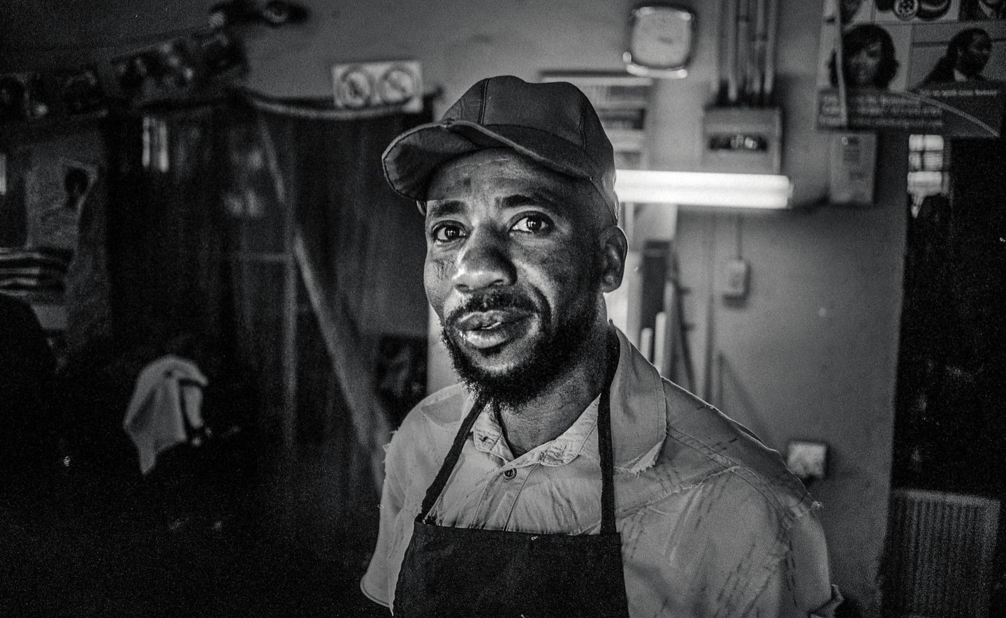 A man wearing a cap and an apron looks directly at the viewer in a dimly lit room.