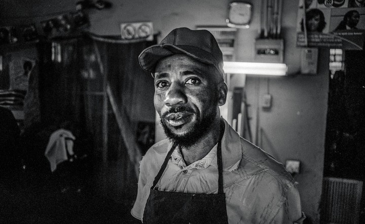 A man wearing a cap and an apron looks directly at the viewer in a dimly lit room.