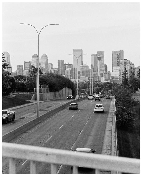 A black and white cityscape shows a highway with cars driving towards a cluster of tall buildings in the distance.