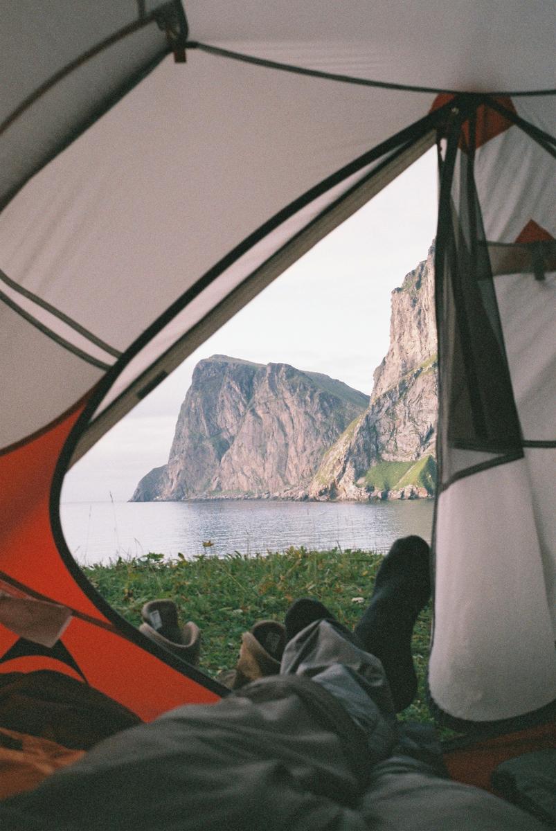 A person's feet are visible inside a tent looking out at a body of water and a large mountain.