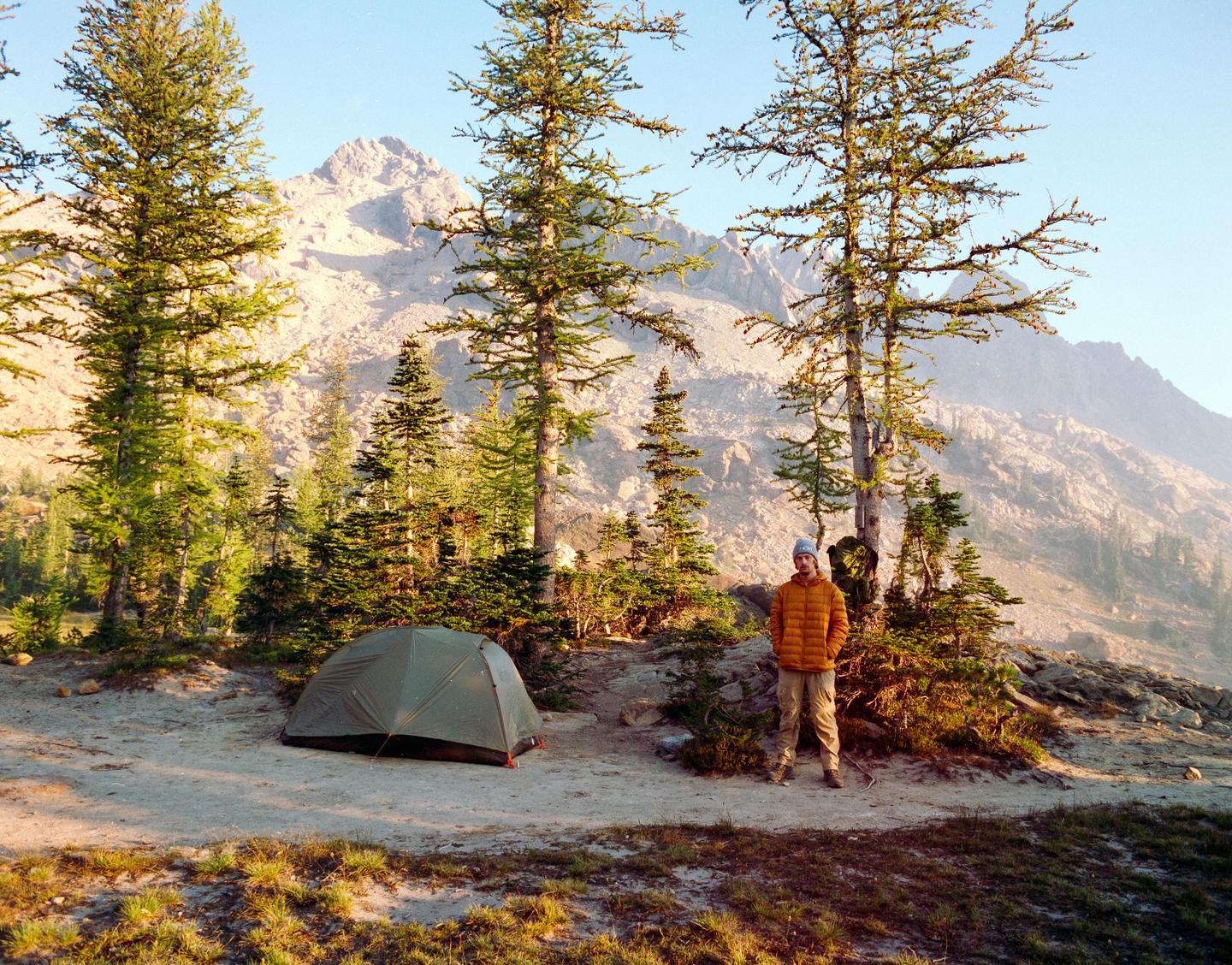 A man stands next to a tent in a mountainous landscape with pine trees.