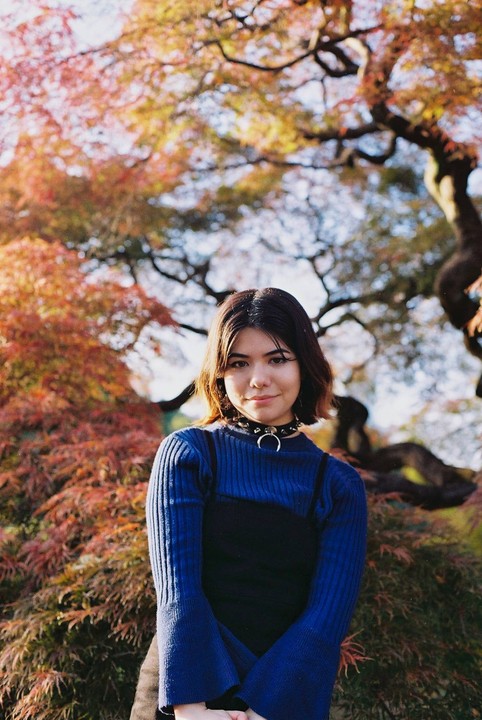 A woman with a choker necklace stands in front of autumn foliage.