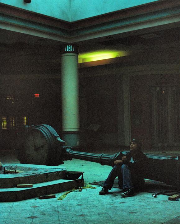 A person sits on the floor next to a fallen lamppost in a dimly lit, abandoned building.