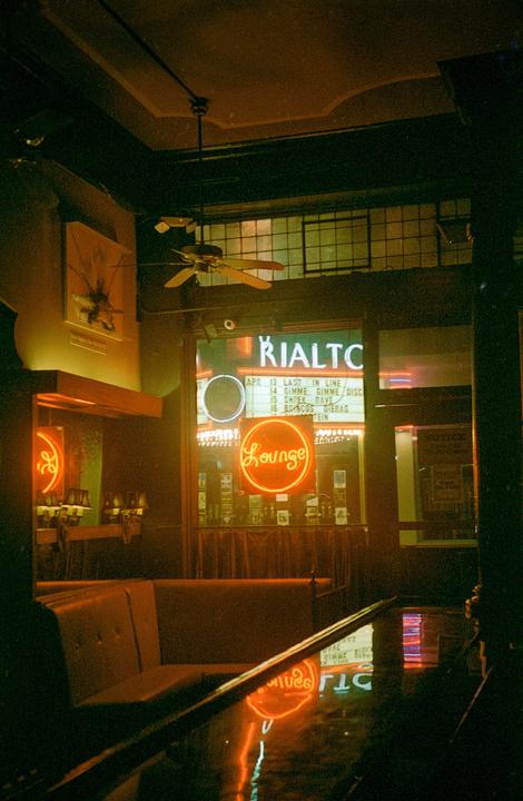 A neon "Lounge" sign is reflected in a wet surface inside a building with a ceiling fan.