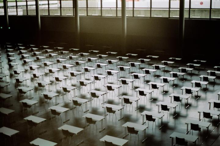 An empty examination hall is filled with rows of desks and chairs, illuminated by light from large windows.