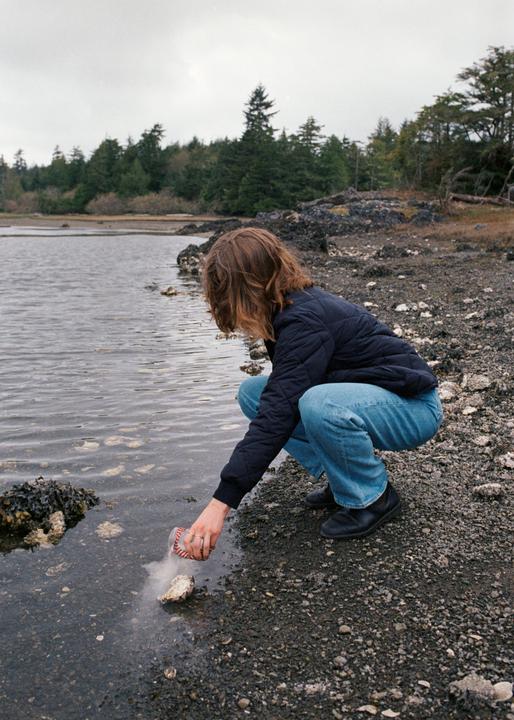 A woman crouches by the water's edge, pouring ashes from a small container onto a rock.