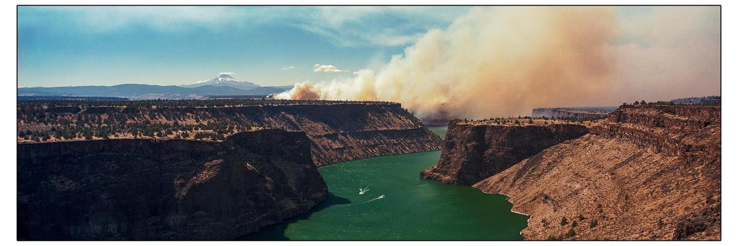 A wide panoramic view shows a river flowing through a canyon with smoke from a wildfire rising in the distance.