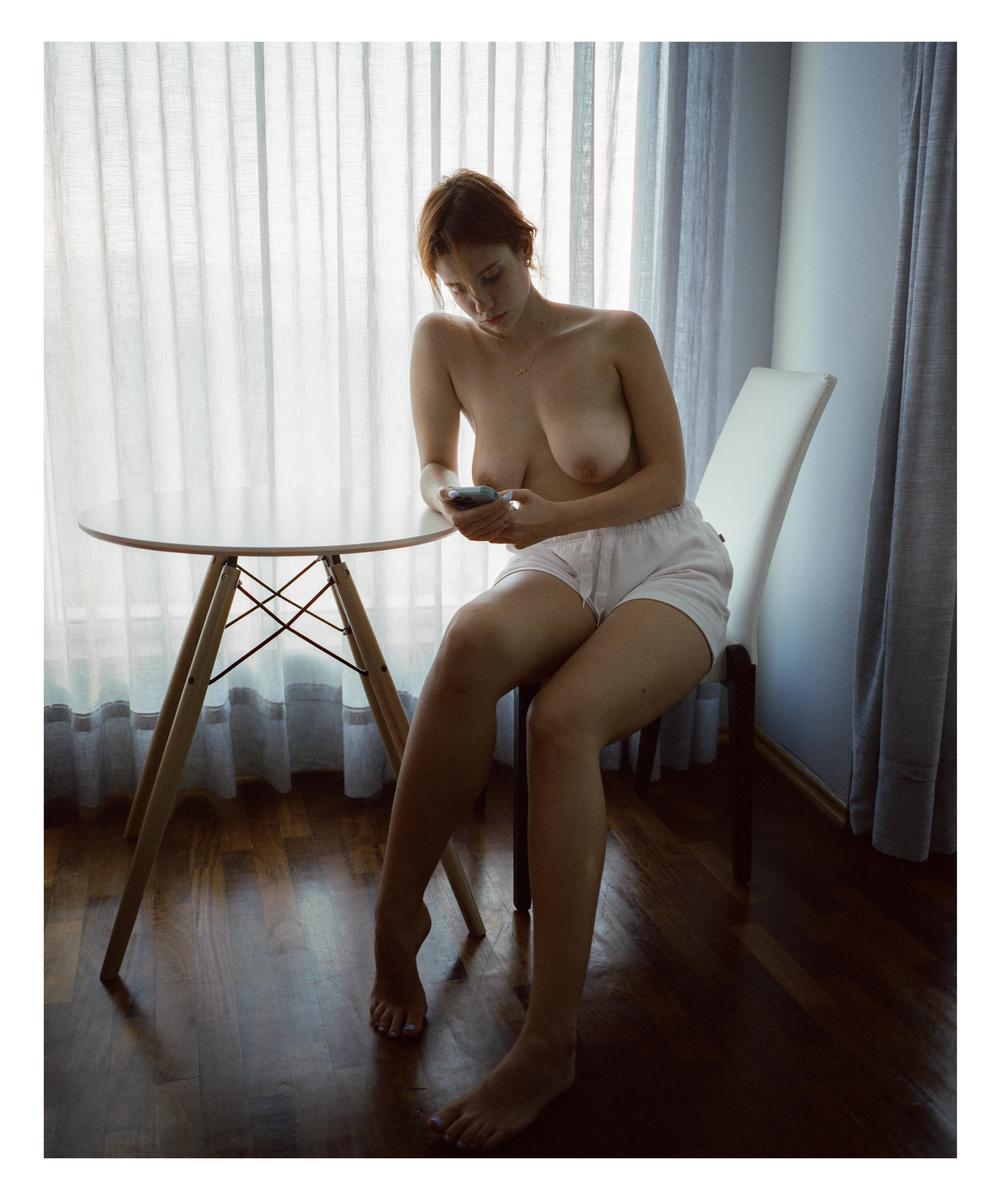 A woman sits topless at a table, looking at her phone.