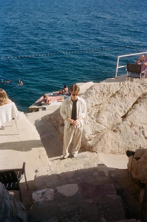 A man stands on a rocky shore overlooking the ocean where people are swimming and sunbathing.
