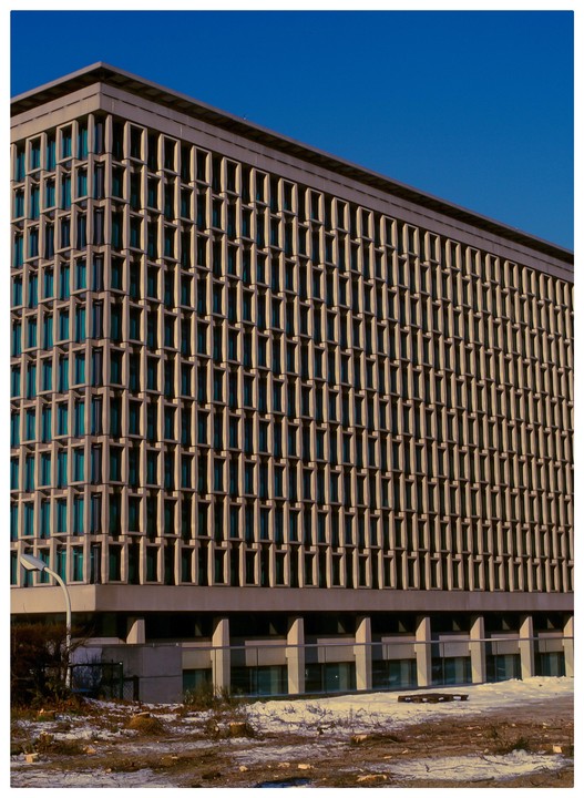 A large building with a grid-like facade stands against a clear blue sky, with a snowy, barren ground in the foreground.