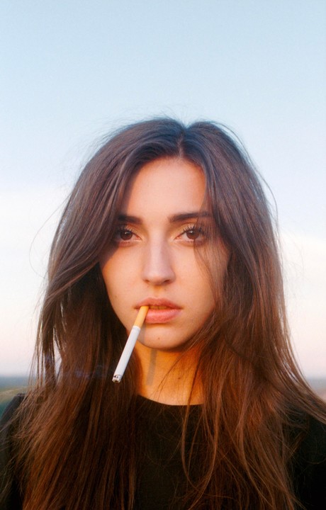 A woman with long brown hair holds an unlit cigarette in her mouth.
