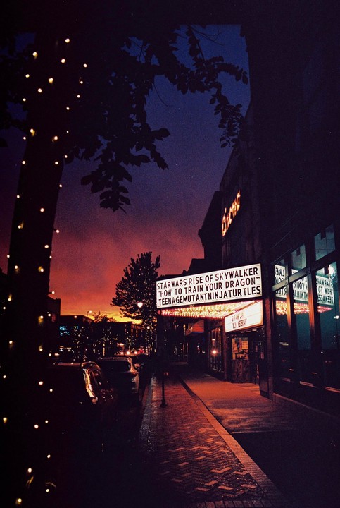 A movie theater marquee displays film titles under a dramatic sunset sky.
