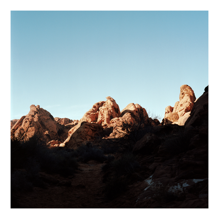 A path leads through a desert landscape with large, sunlit rock formations under a clear sky.