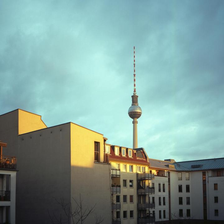 The Berlin Fernsehturm rises above buildings with sunlit walls under a cloudy sky.