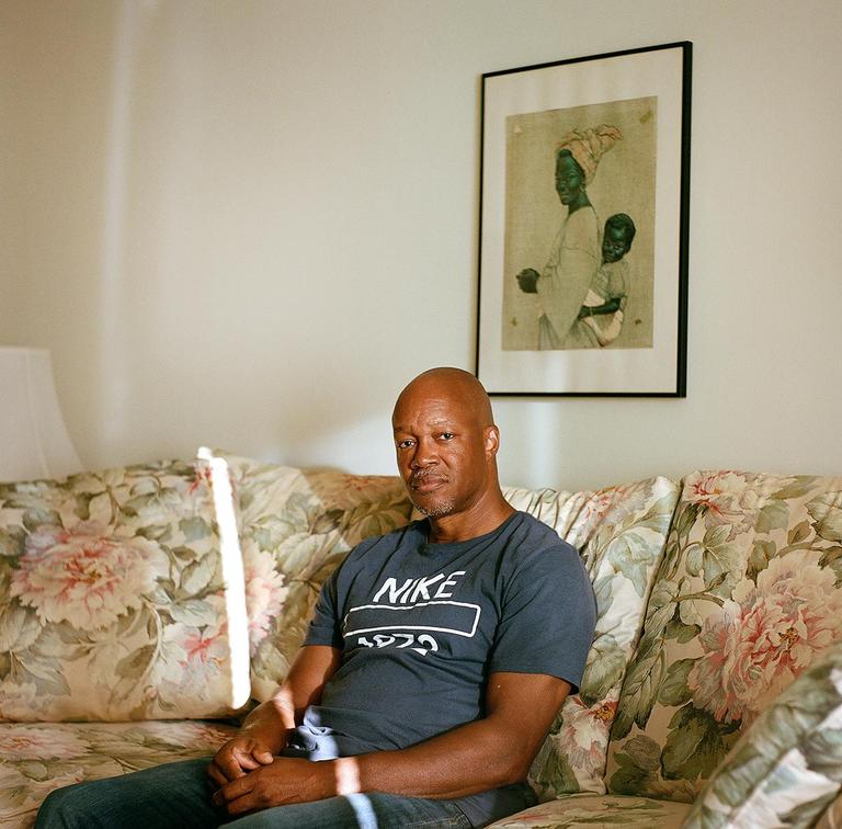 A man sits on a floral couch with a framed picture of a woman and child on the wall behind him.