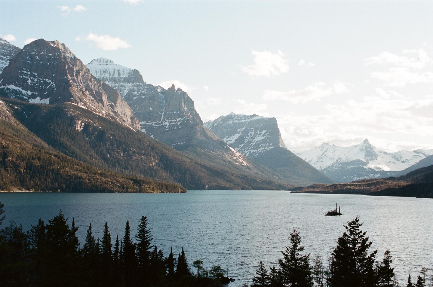 A wide shot shows a lake surrounded by mountains with snow-capped peaks and a small island with trees and a structure.