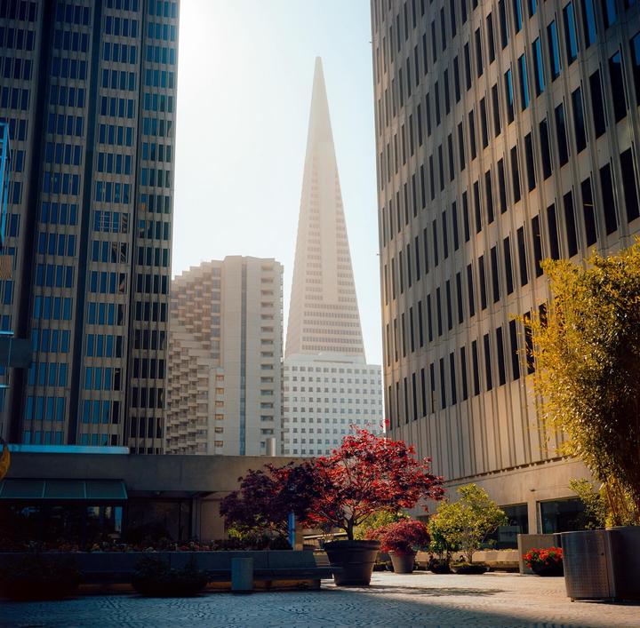 A tall, pointed skyscraper rises above other buildings in a city square with trees and planters.