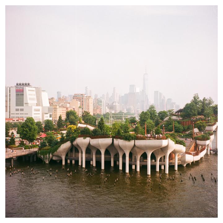 A modern park structure with trees and people on a body of water is visible in the foreground, with a hazy city skyline in the background.