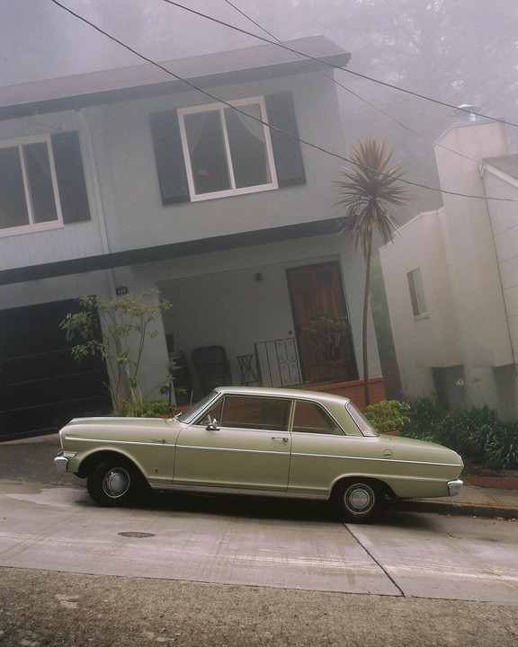 A light green car is parked on a street in front of a house on a foggy day.