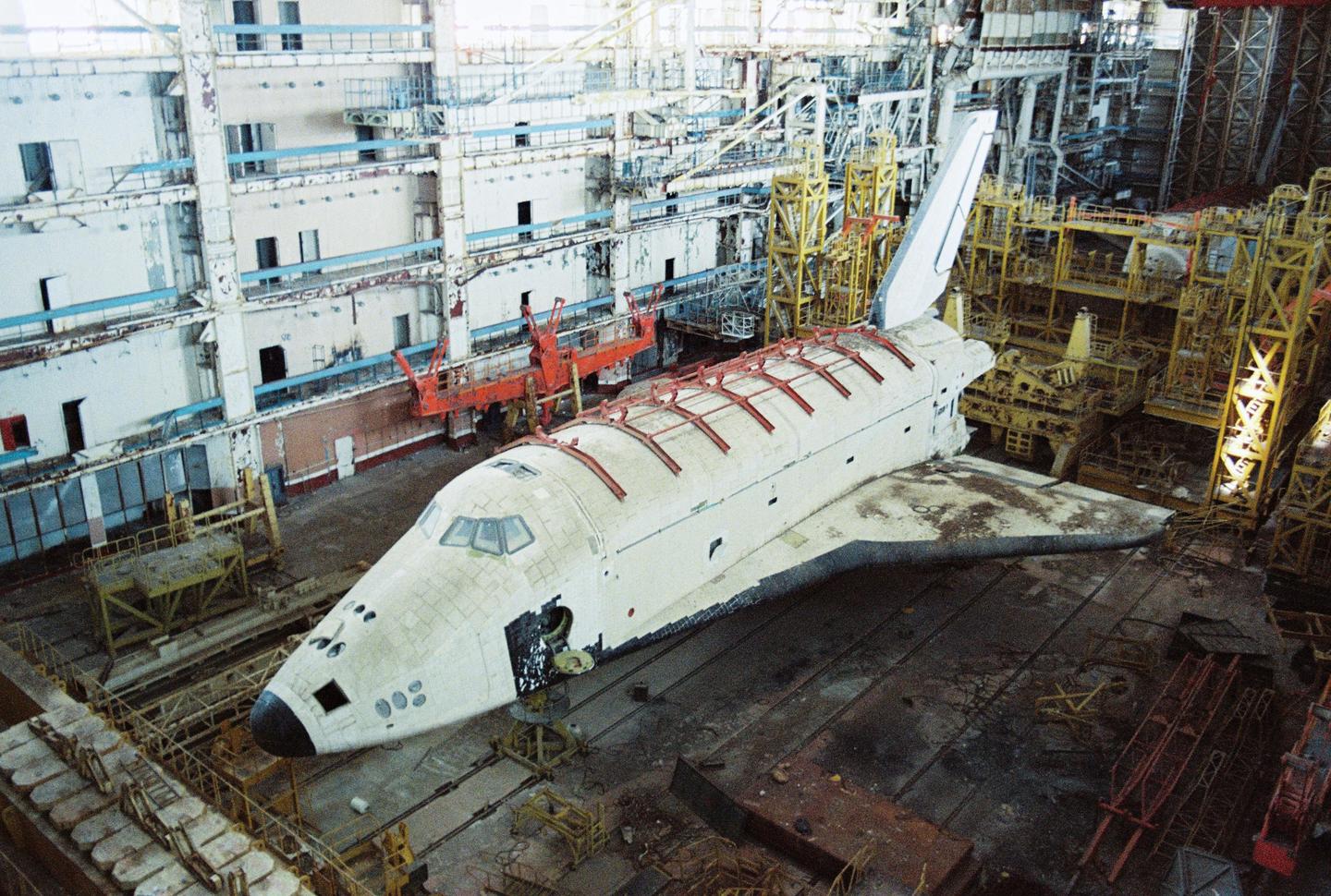 A large, white, abandoned space shuttle sits in a hangar at the Baikonur Cosmodrome.