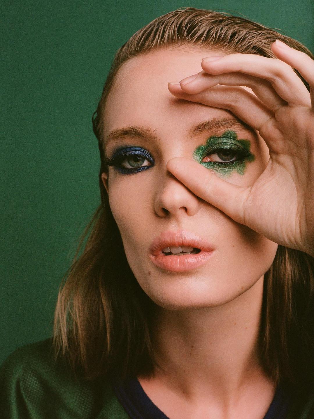 A woman with blue and green eyeshadow covers part of her face with her hand.