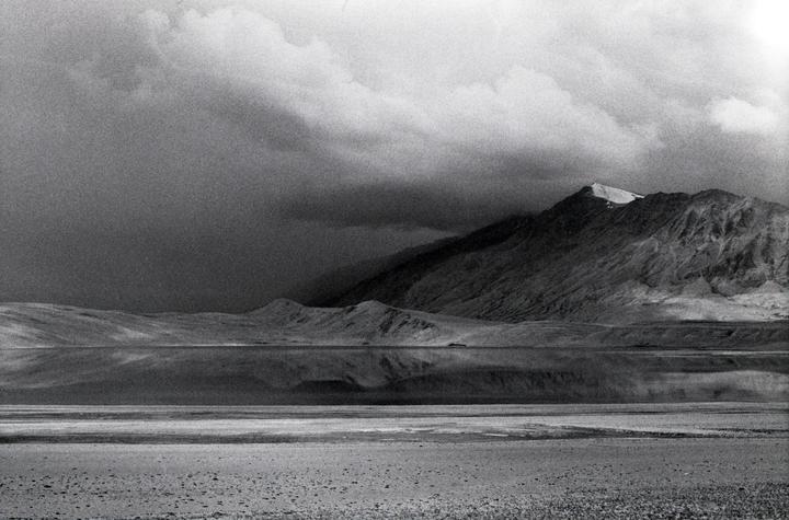 A mountain range with a snow-capped peak is reflected in a still body of water under a cloudy sky.