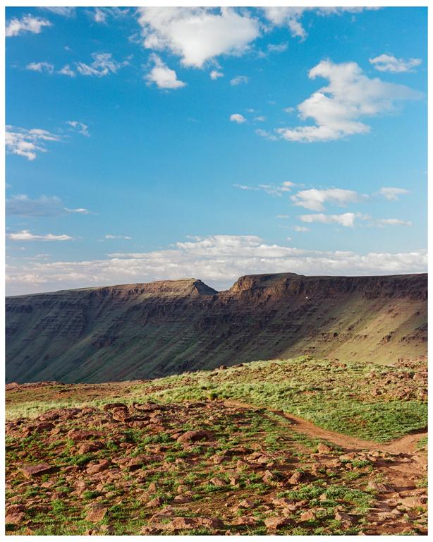 A dirt path leads through a rocky, grassy landscape towards a large mountain under a blue sky with clouds.