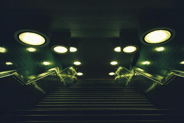 A long, dark staircase with bright, circular lights on the ceiling and walls leads upwards.