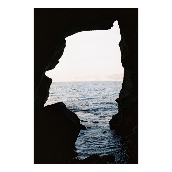 A dark cave opening frames a view of the ocean and a distant coastline under a bright sky.