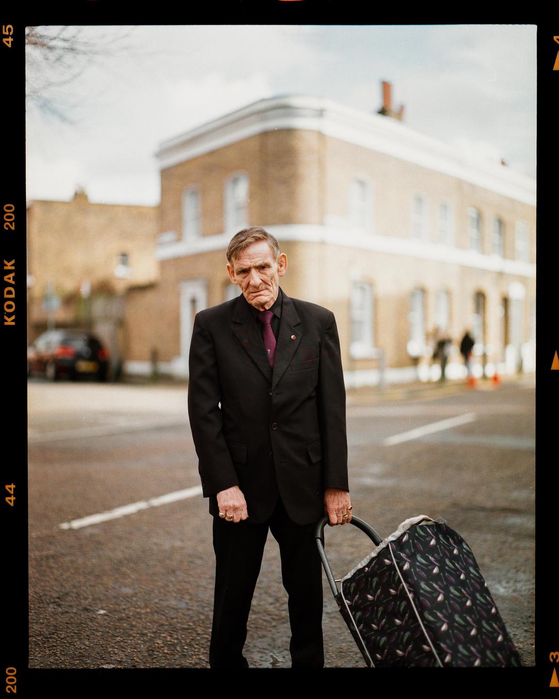 A man in a suit stands on a street holding a shopping trolley.