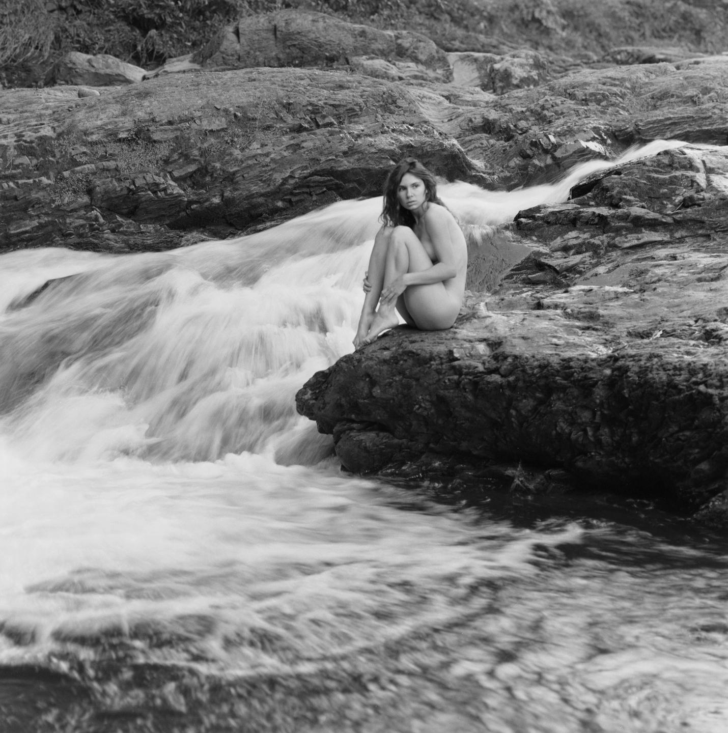 A woman sits on a rock next to a flowing river.