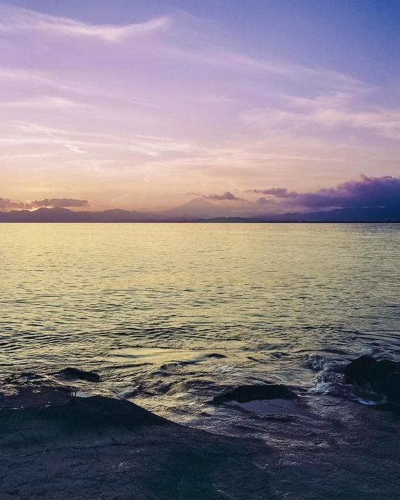 A body of water with rocks in the foreground reflects the colorful sky at sunset, with mountains visible in the distance.