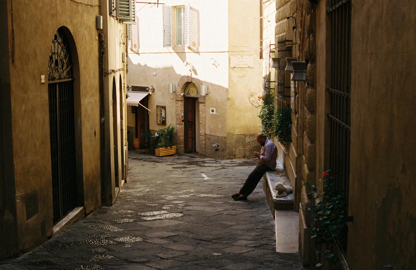 A man sits on a stone bench with a dog next to him in a narrow alleyway.