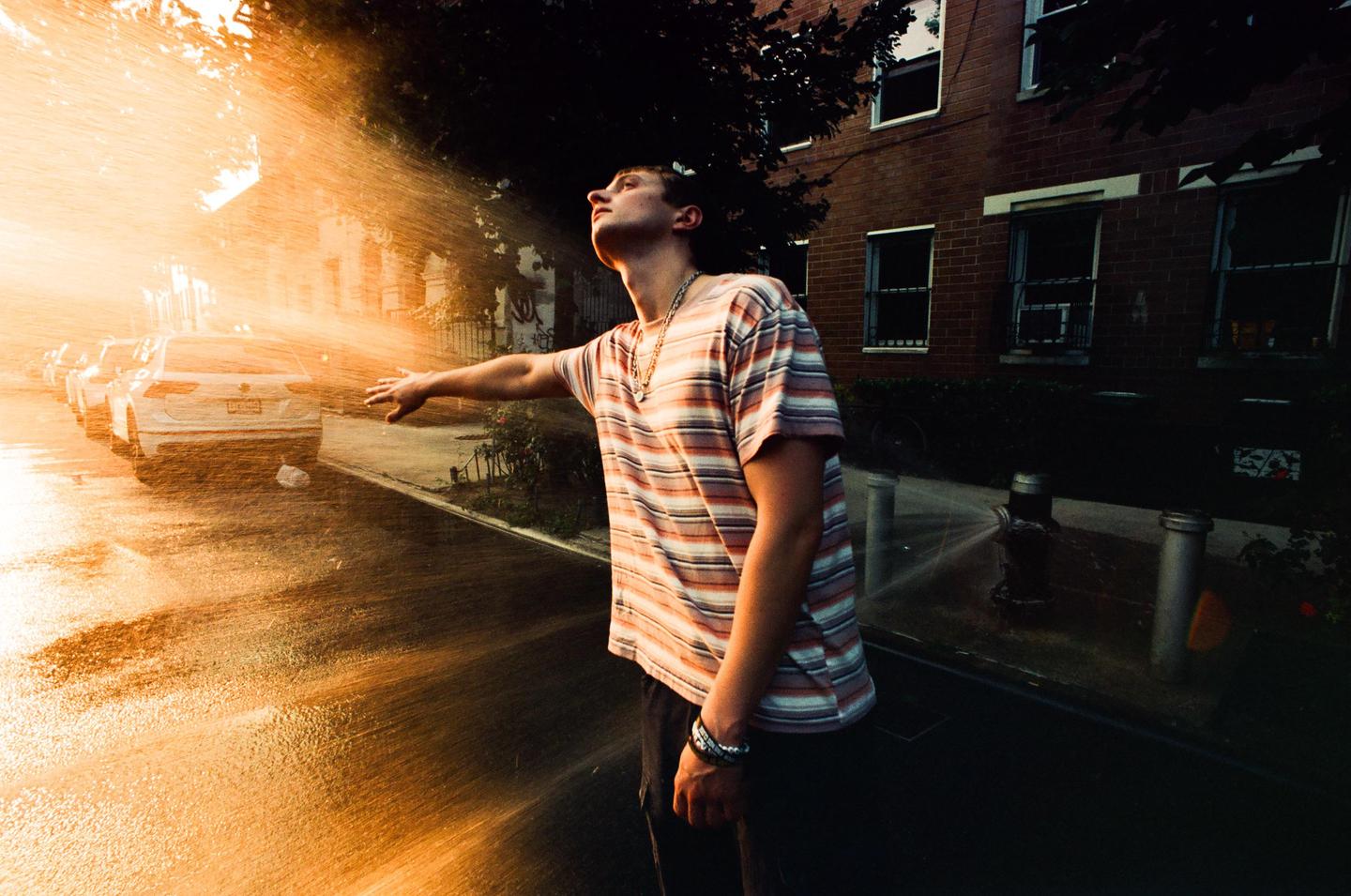A man stands on a street with his arm outstretched into the spray from a fire hydrant, with cars and a brick building in the background.