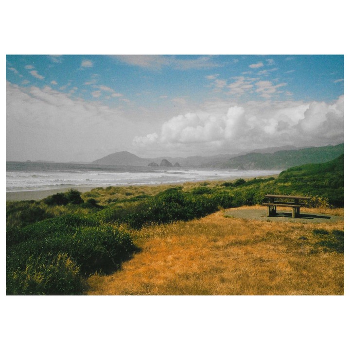 A picnic table sits on a grassy hill overlooking a beach with waves crashing, mountains in the distance, and a cloudy sky.
