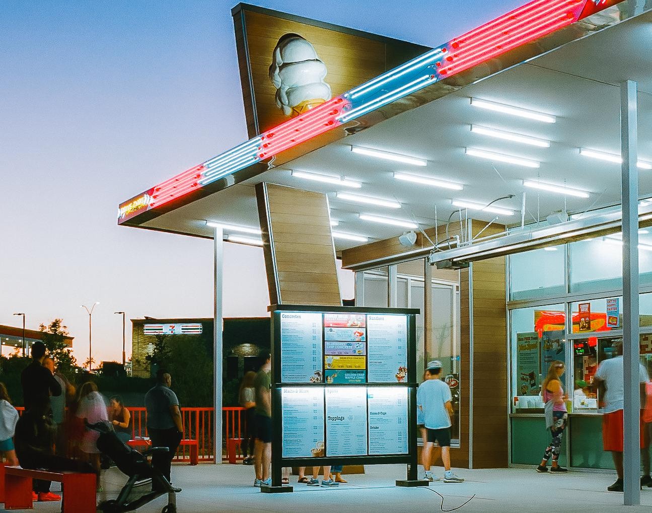 People gather at an outdoor ice cream stand with neon lights at dusk.